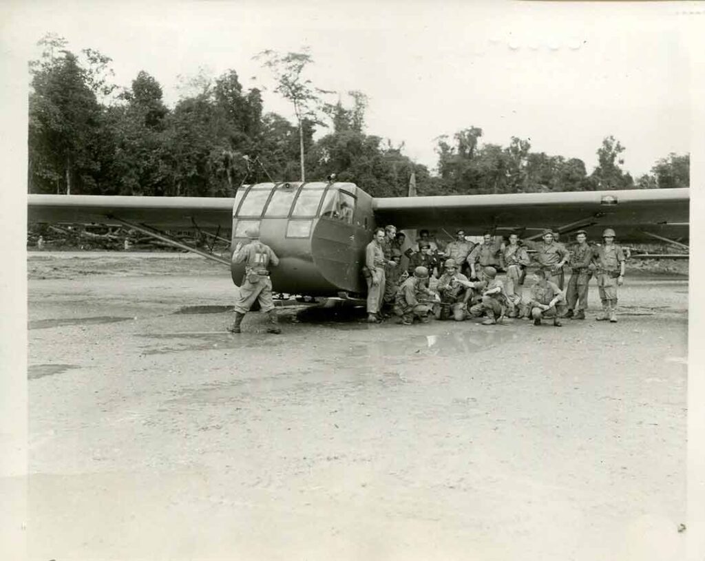 Glider pilots and airborne engineers stand in the shade of a glider wing at an airfield in northern Burma as they wait for H-Hour for the assault on the Japanese-held airfield at Myitkyina, 17 May 1944. (National Archives)